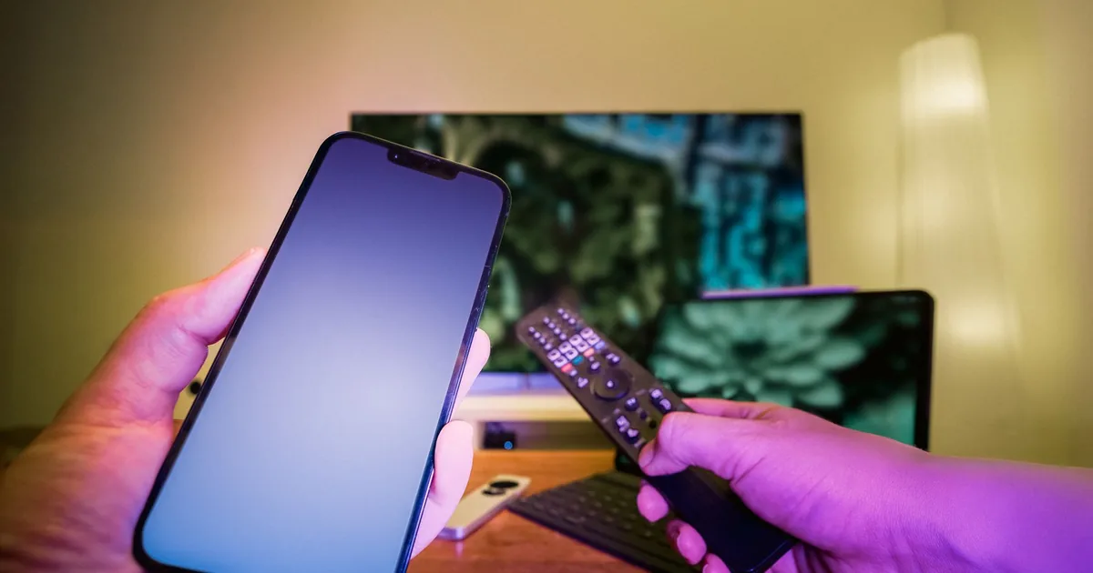 Hands holding a smartphone and remote control in front of a TV and home entertainment devices