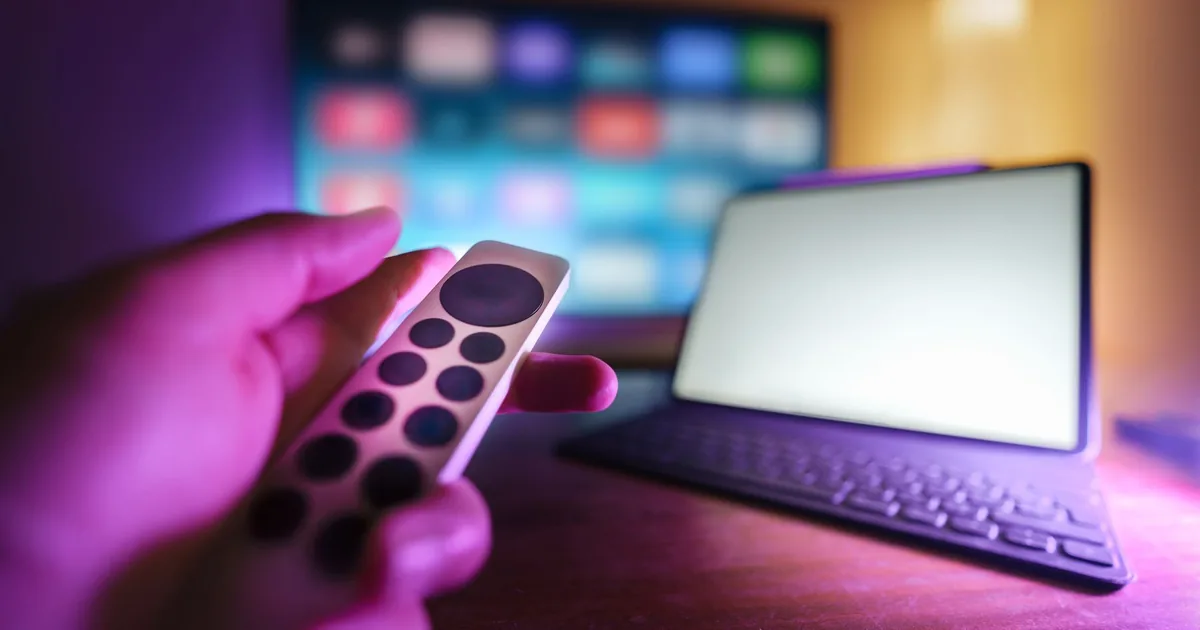 Hand using a remote control with a TV and laptop in a cozy room
